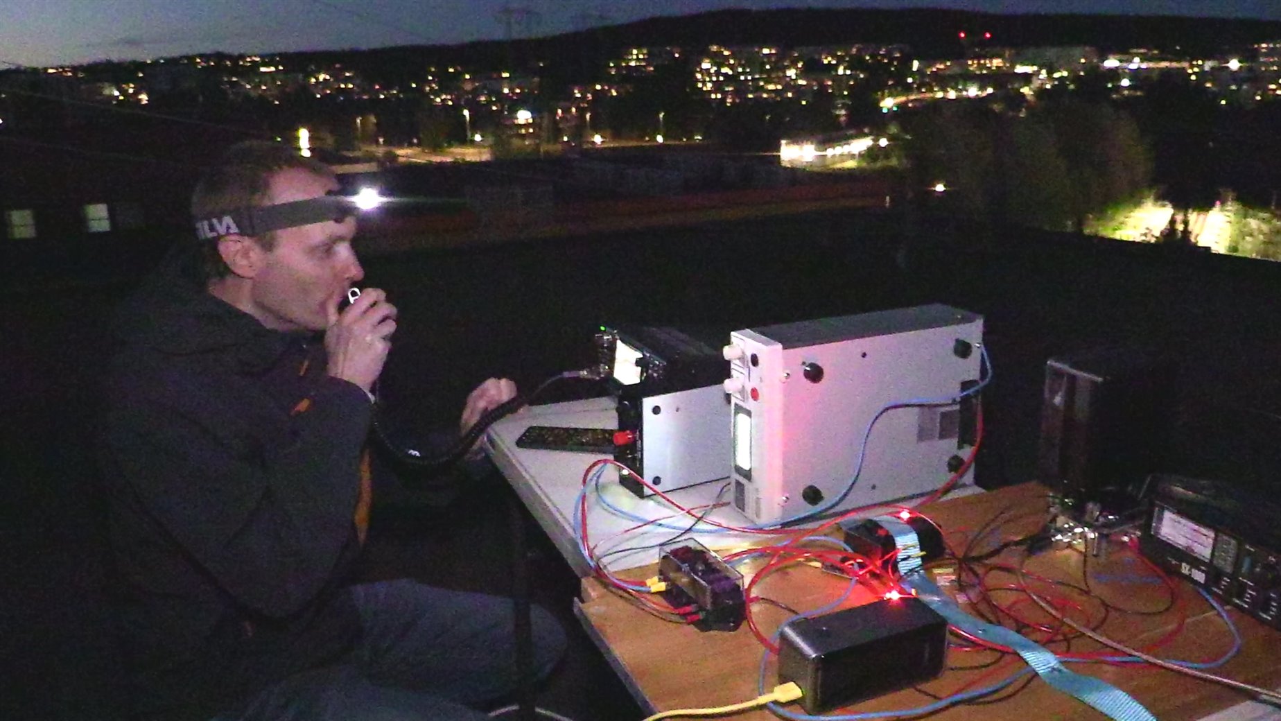 An operator wearing a headlamp speaks into a microphone at a folding table of radio equipment on a rooftop at night, with the lights of Oslo behind.